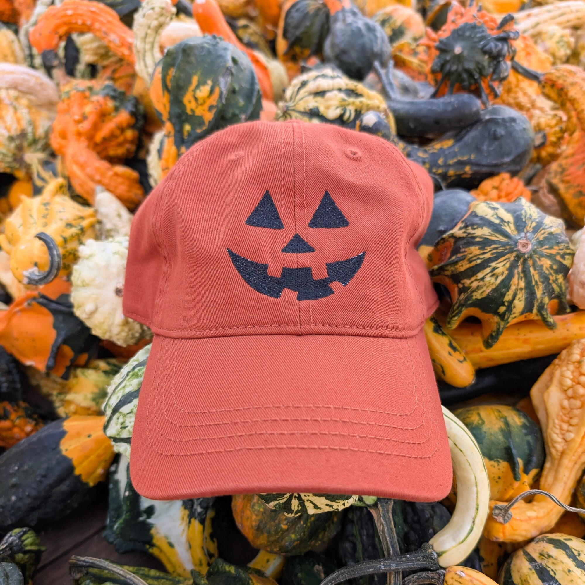 Red baseball cap with a jack-o'-lantern face, surrounded by various gourds and pumpkins, Halloween-themed display.