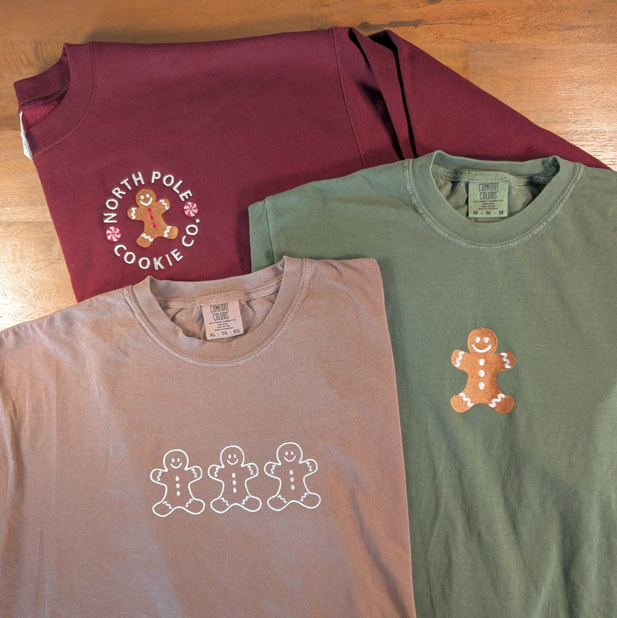 Three T-shirts with gingerbread man designs, one maroon with logo, one beige with three gingerbread men, one green with a single gingerbread man, on a wooden surface.