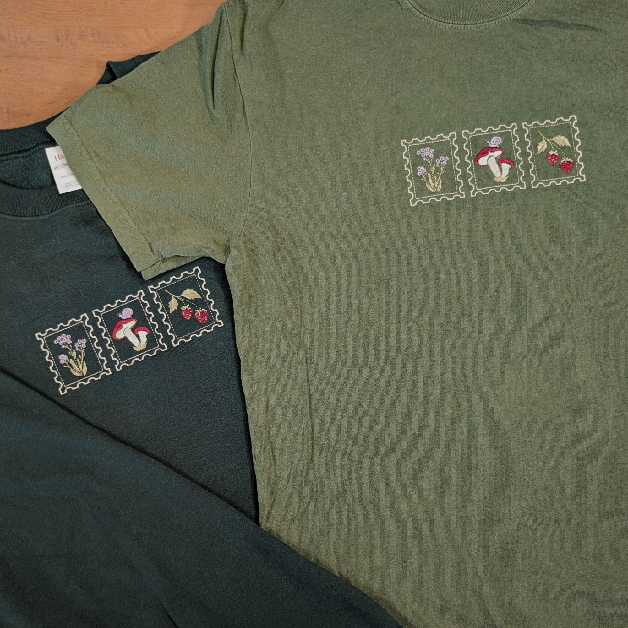 Two T-shirts with embroidered postage stamp designs featuring animals and a person, one green and one black, laid flat.
