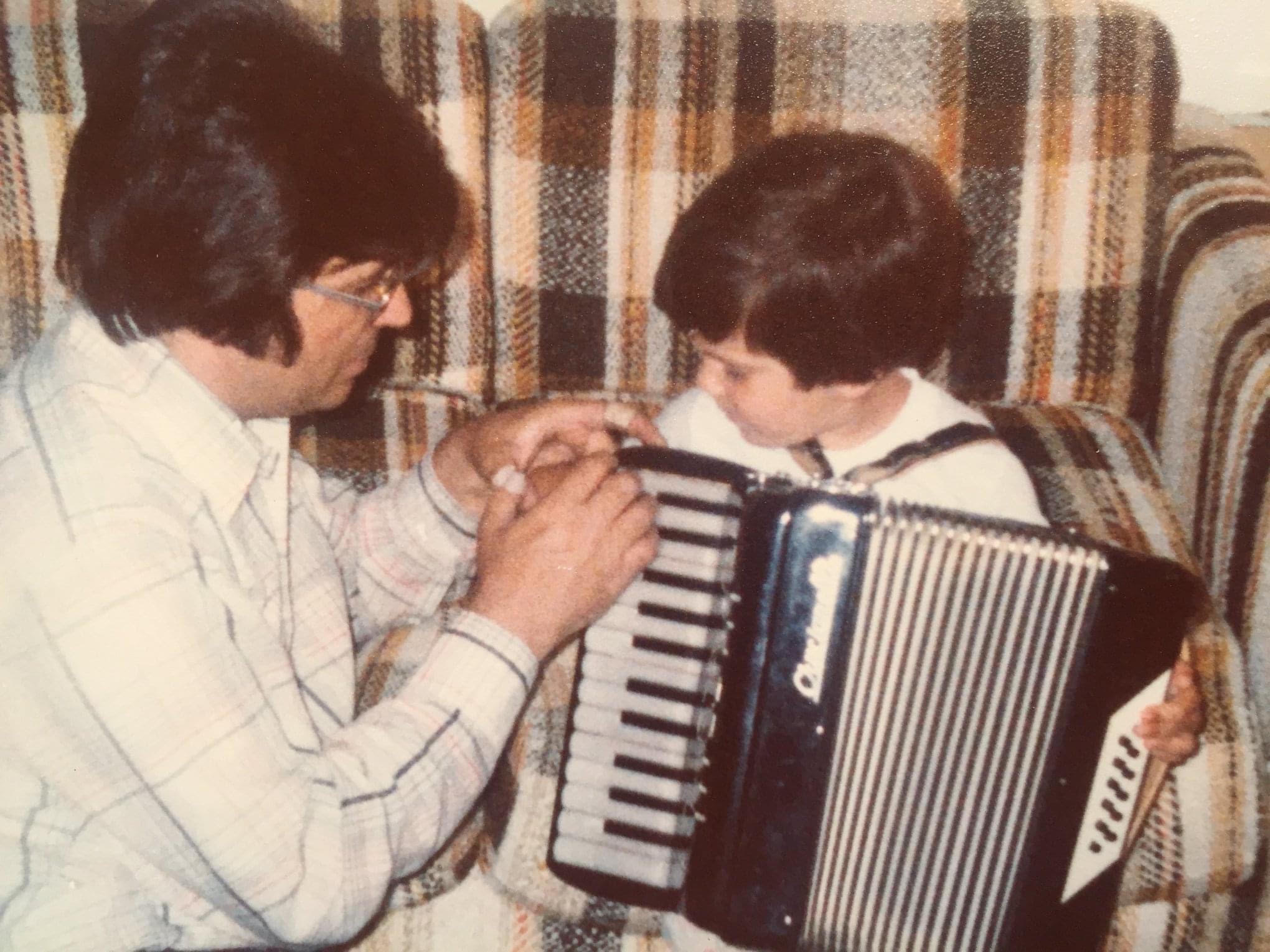 Adult and child sitting on a plaid sofa, with the adult helping the child play an accordion.