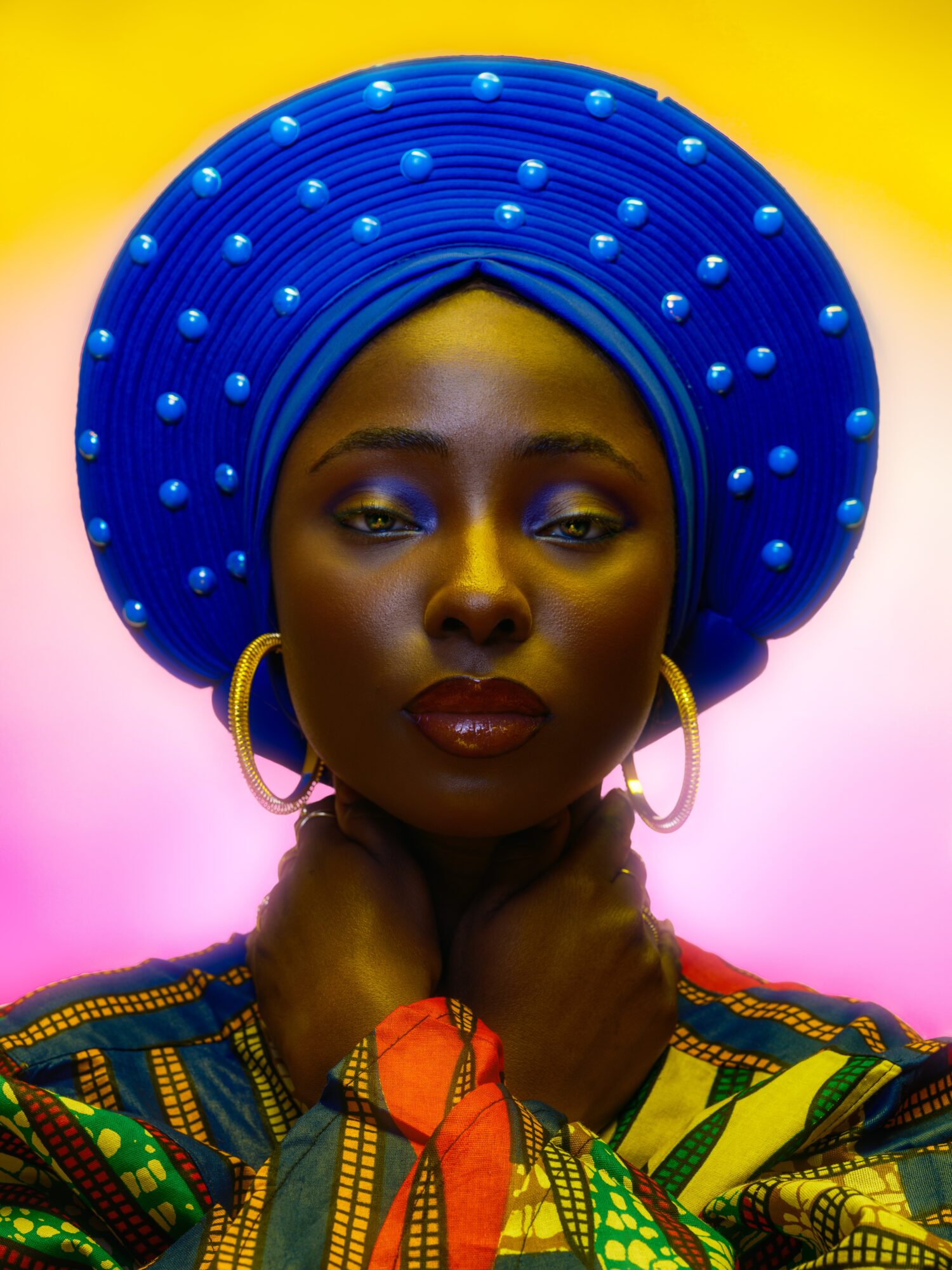 Portrait of a woman wearing a large blue headwrap with pearls, gold hoop earrings, and colorful clothing, against a vibrant background.