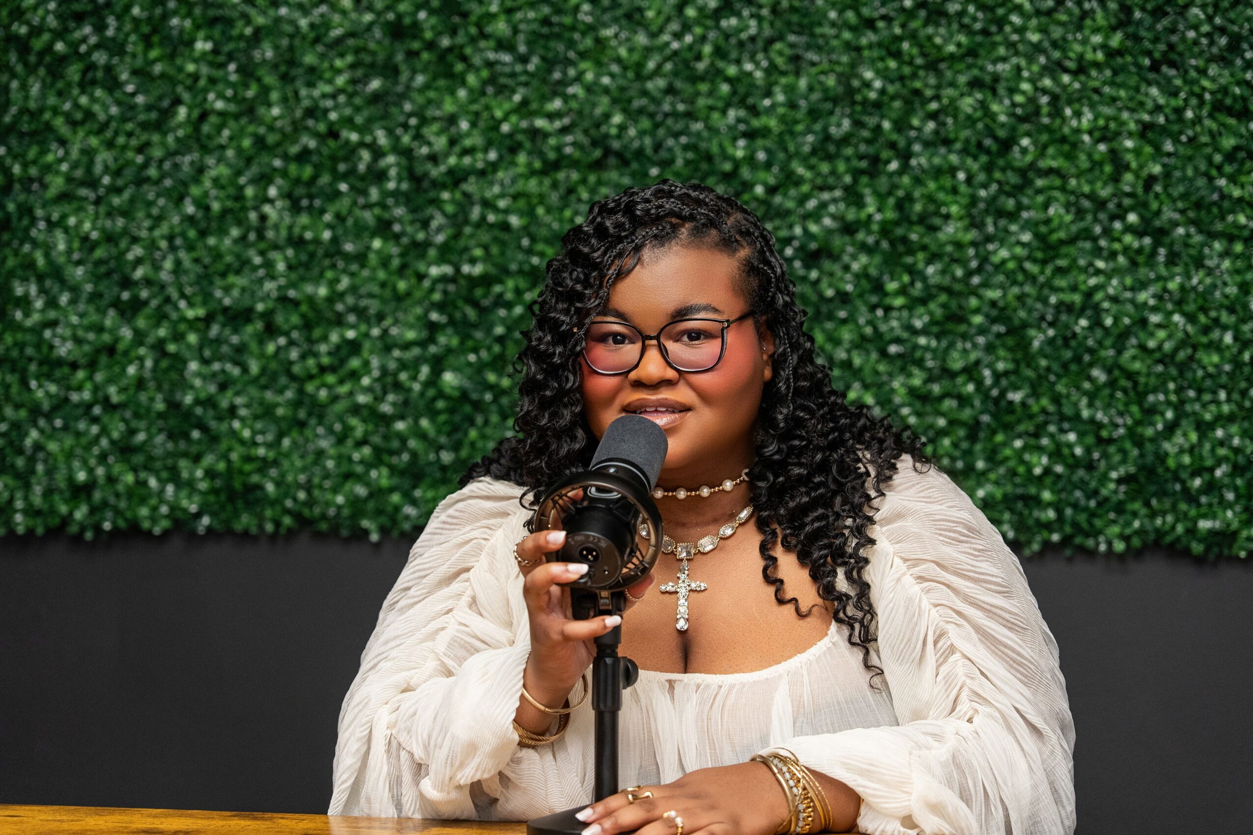 Woman with curly hair and glasses speaking into a microphone, wearing a white outfit and jewelry, standing in front of green foliage