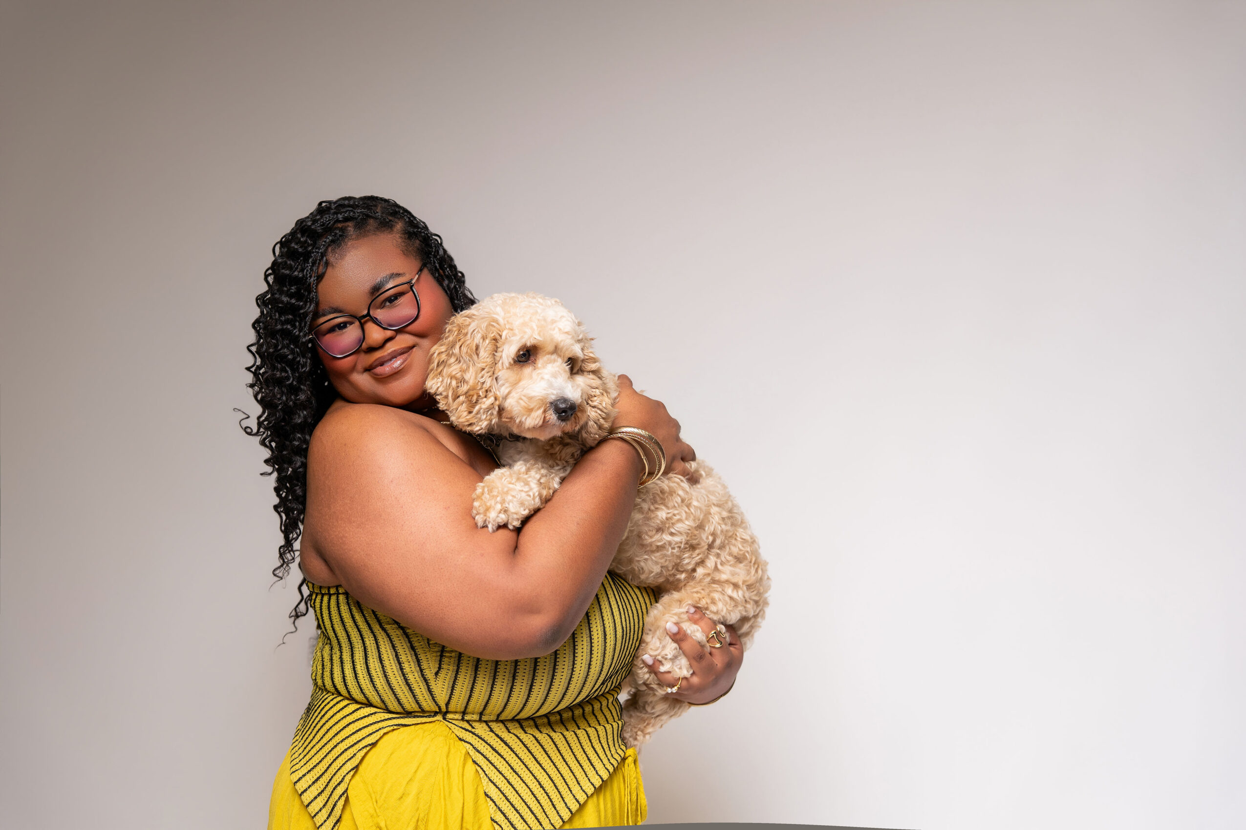 A woman with curly hair and glasses holding a fluffy dog against a plain background.