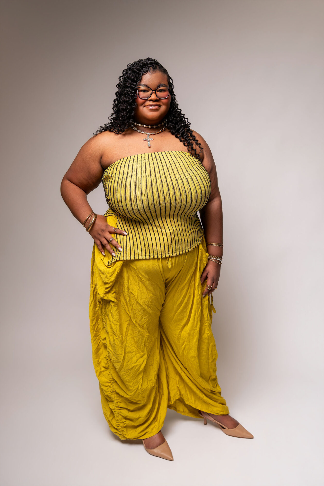 Woman with curly hair and glasses wearing a yellow strapless top and matching wide-leg pants, standing with one hand on her hip.