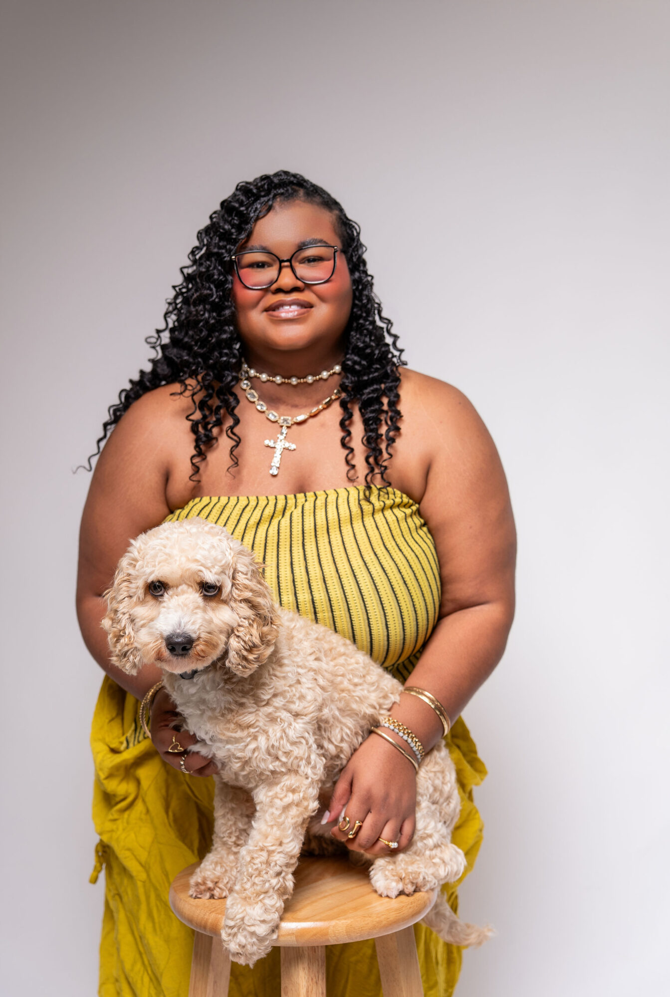 Smiling woman with curly hair, glasses, and jewelry holds a small fluffy dog on a stool against a plain background.