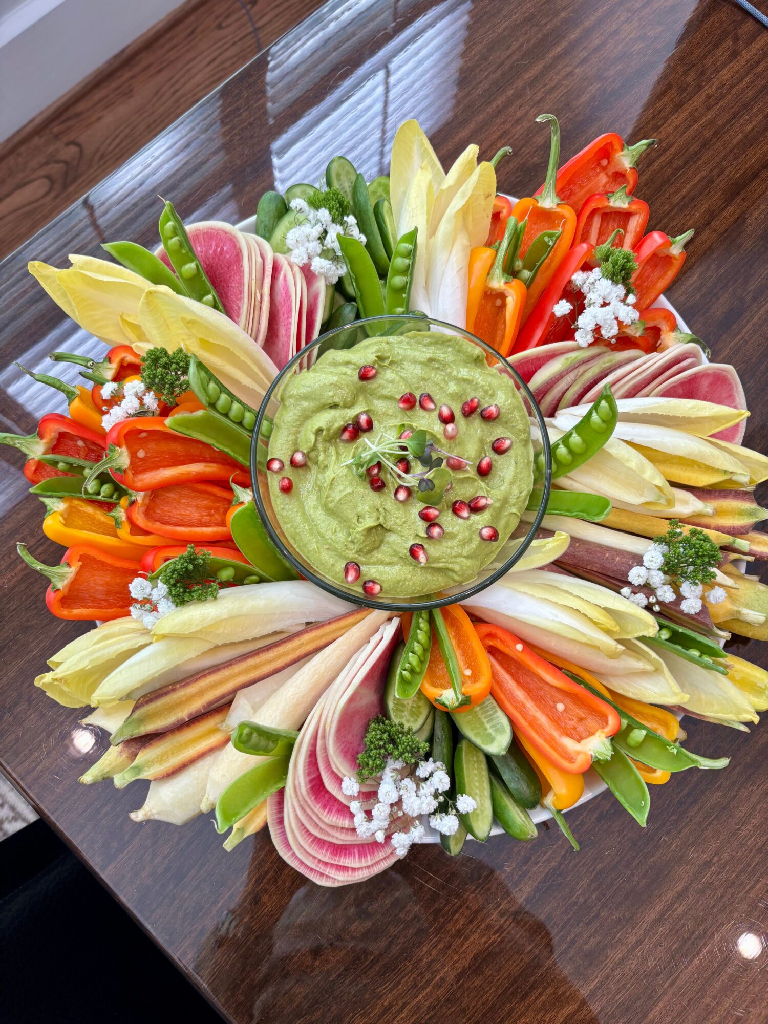 Colorful vegetable platter with a central green dip, surrounded by sliced vegetables like carrots, radishes, and cucumbers, garnished with herbs and flowers.